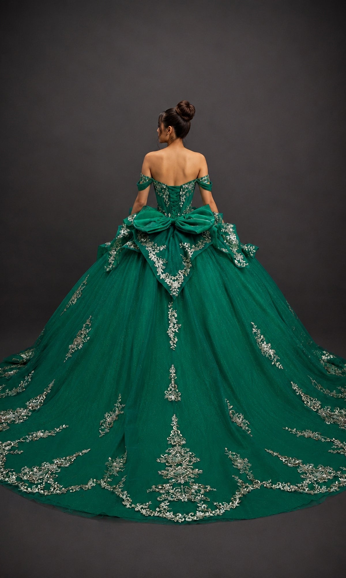 A woman stands facing away against a dark background, wearing the Quinceanera Dress 5198 By Dancing Queen—an off-the-shoulder green ball gown adorned with silver embroidery and featuring a fitted corset bodice.